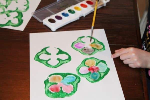 Easy Preschool Shamrock Craft - child painting watercolor rainbow colors into a 4 leaf clover shape.