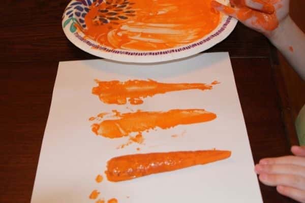 child making carrot stamps by dipping a carrot half into a plate of orange paint and stamping it onto a white piece of paper