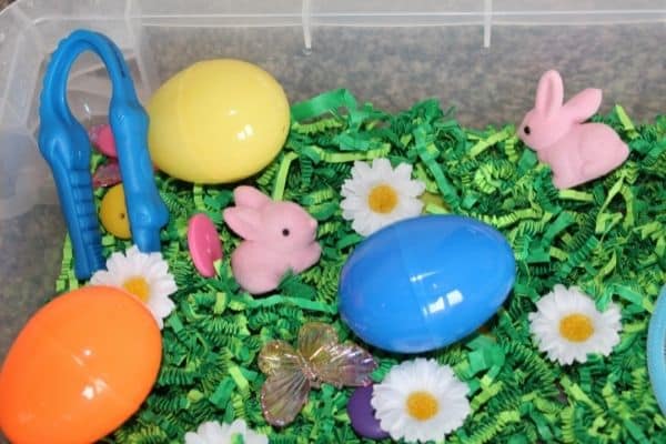 Easter sensory bin with plastic eggs, tongs, bunnies, flowers, and butterfly.