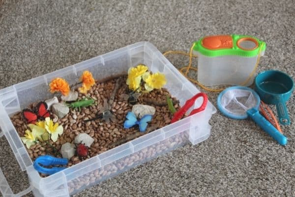 Insect sensory bin with beans, fake bugs and insects, a mini butterfly net, a scoop, a mini bug collector, stones, sticks, tongs, and more.