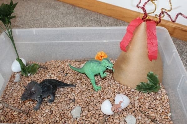 Dinosaur sensory bin filled with beans, dinos, eggs, and paper volcano