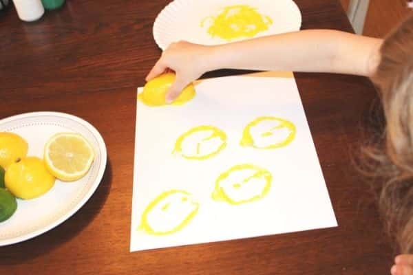 Child stamping yellow lemon prints onto a white paper next to a plate of lemons