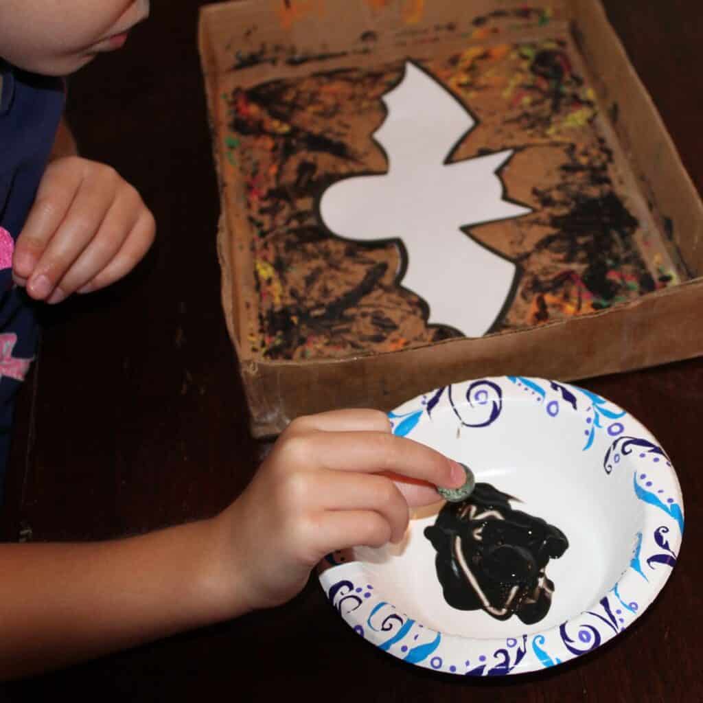 bat template in box, child is dipping marble into black paint to prepare to marble paint