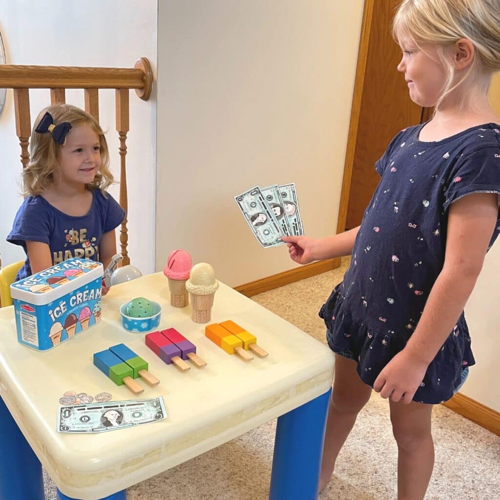 Kids playing ice cream shop. One girl is paying fake money while the other serves her ice cream