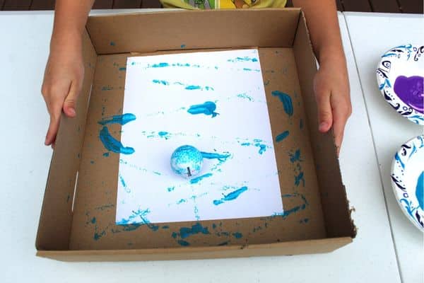 child shown painting with a golf ball in a box - painting with balls activity for preschool