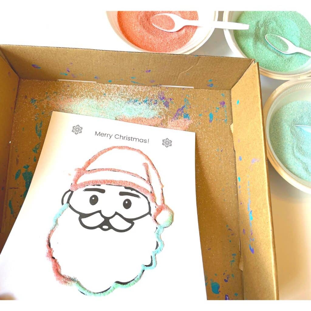 Santa preschool craft sand art project for kids - shown Santa face printable with glue and sand - extra sand being shaken off into a shallow box