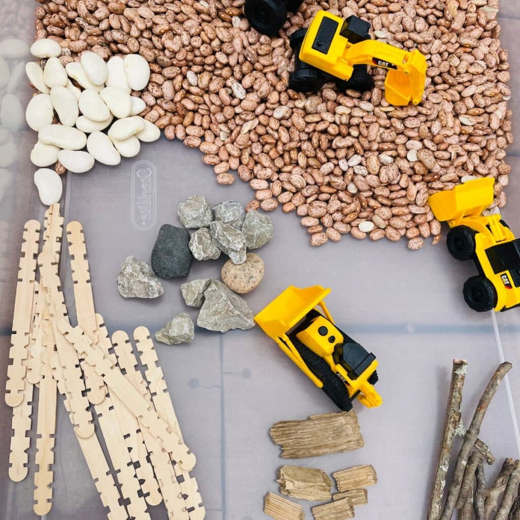construction sensory bin - a construction themed sensory activity - contains construction vehicles, beans, rocks, sticks, sawtooth popsicle sticks, lima beans and wood chips