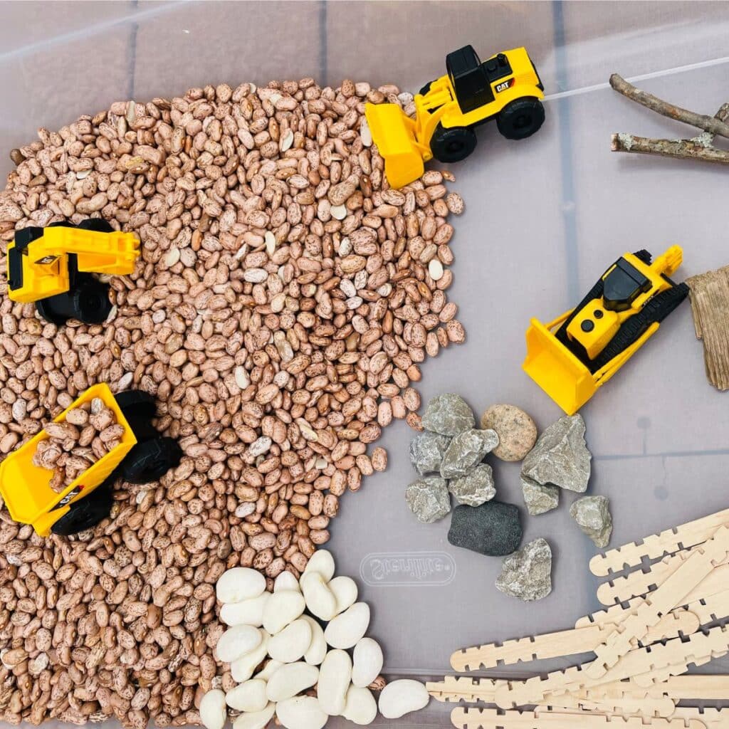 Construction sensory bin - a construction themed sensory play idea for preschool - construction vehicles hauling pinto beans, lima beans, rocks, sticks, wood chips, and popsicle sticks.