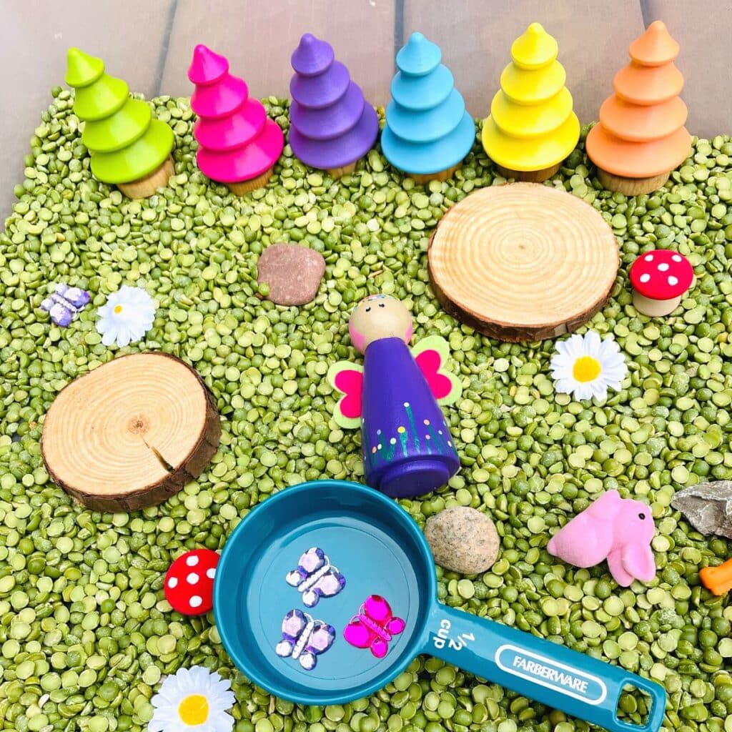 fairy sensory bin - a fairy garden sensory play idea shown: wooden fairy with painted rainbow wooden trees, cups to scoop and more.