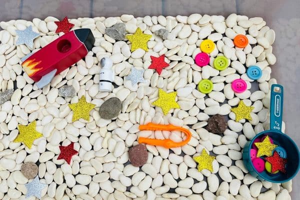 outer space sensory bin - a fun space sensory play idea including wooden astronaut and rocket, lima beans, tweezers, rocks, buttons, a scoop, and foam stars