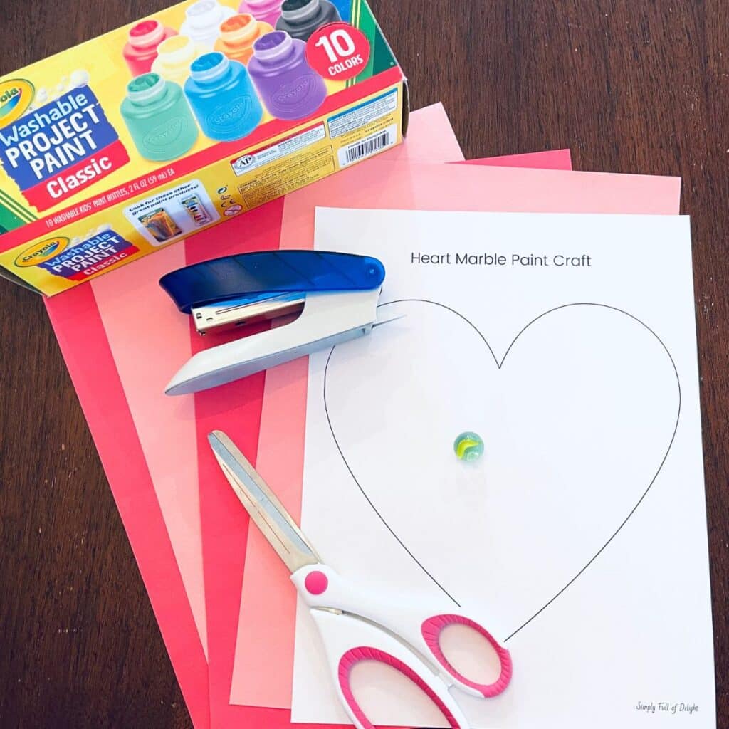 Supplies for Valentine's Day marble painting craft include paint, free heart printable, marble, stapler, scissors, and colored paper