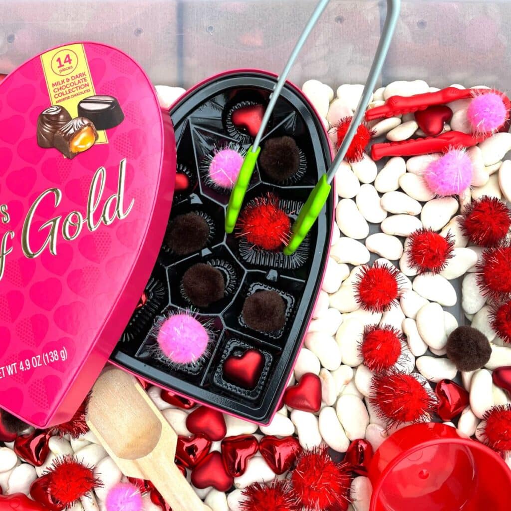 Valentine's Day sensory bin - close up of tongs with pom poms and a chocolate box