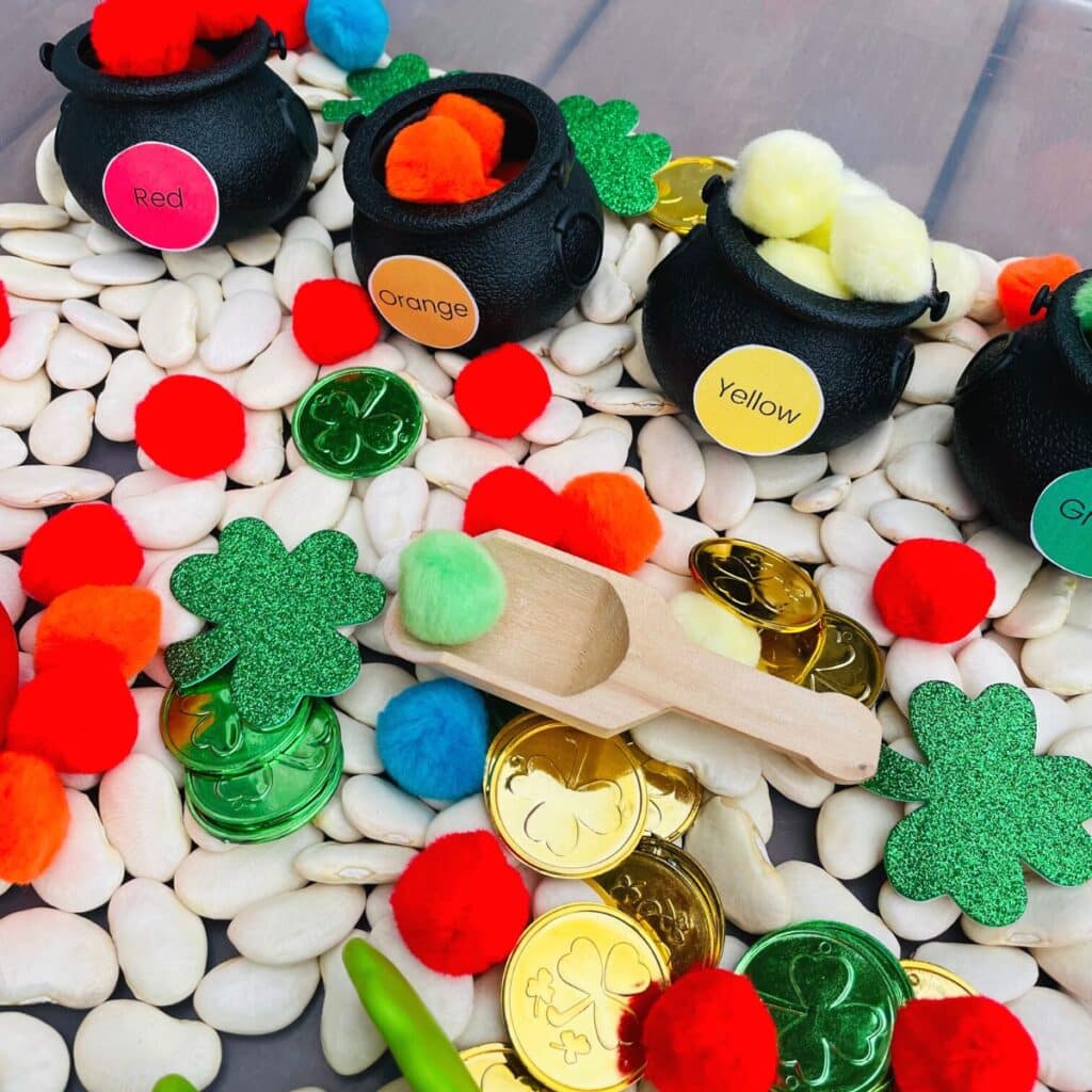 St. Patrick's Day sensory bin for preschool - lima beans, wood scoop, pom poms, shamrocks, gold coins, and pots of gold