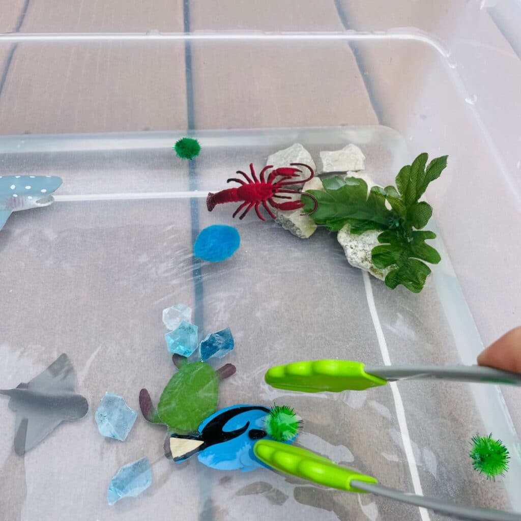 using tongs to pick up a pom pom in an ocean sensory bin