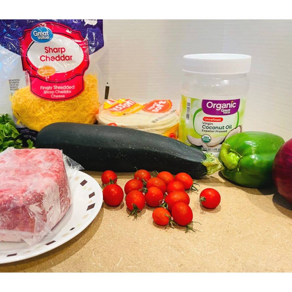 ingredients for zucchini tacos - shown: ground beef, green bell pepper, red onion, cherry tomatoes, coconut oil, mission soft corn tortillas, zucchini, cilantro, and shredded cheddar cheese